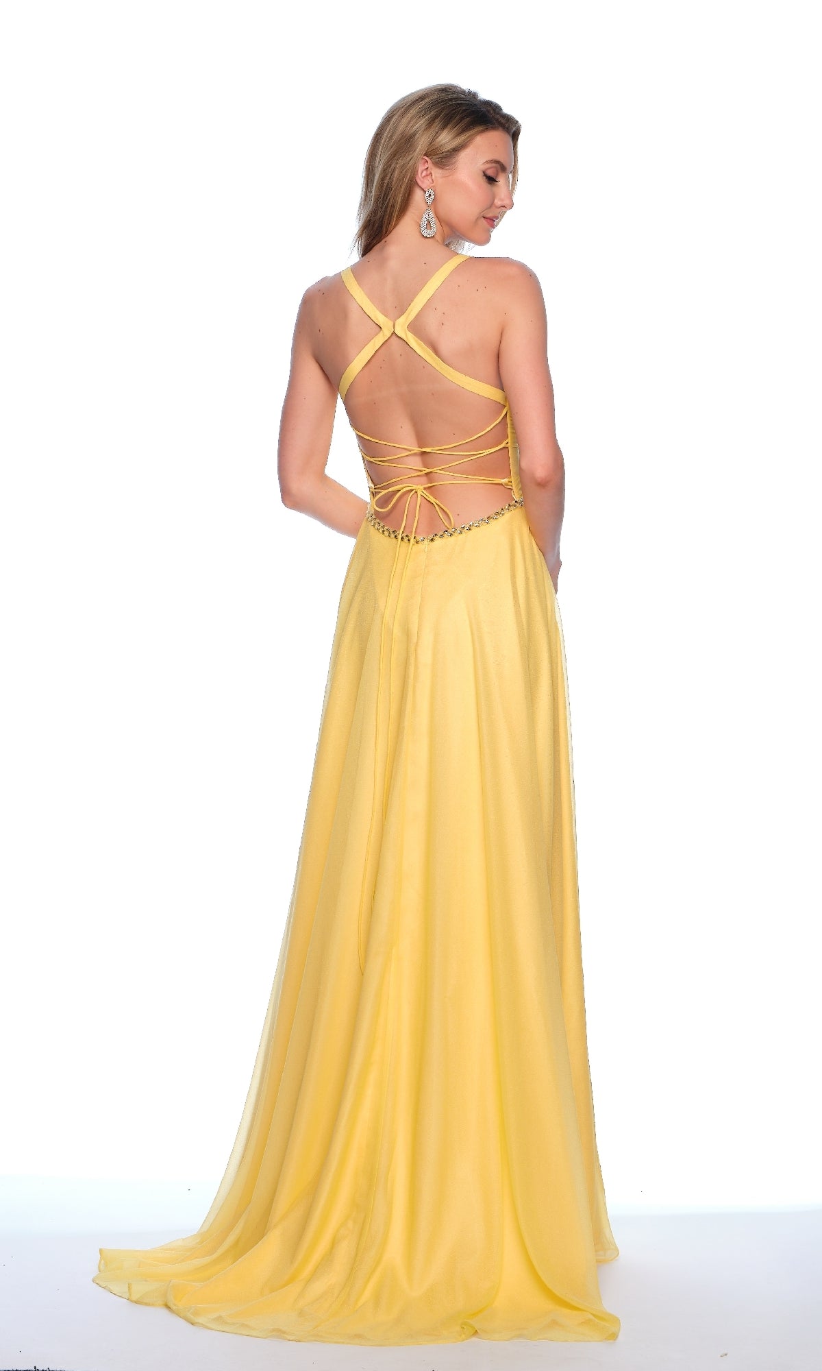A woman stands with her back to the camera in the Dave & Johnny 11872 long prom dress—a sleeveless yellow gown with a v-neck bodice, crisscross open-back straps, and a flowing skirt, set against a white background.